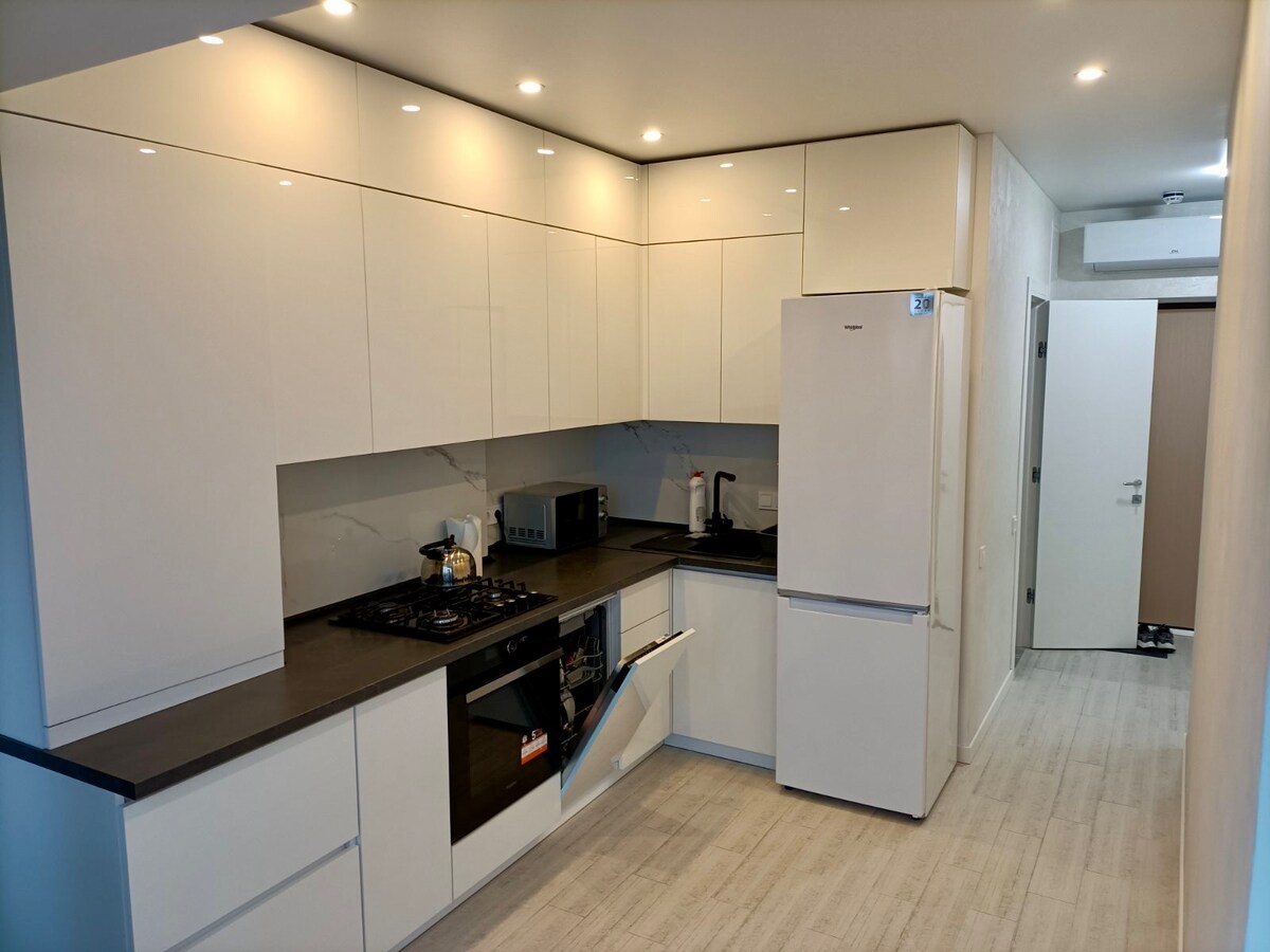 The kitchen features modern white cabinetry with sleek finishes and ample storage. A gas stove and a refrigerator are prominently displayed. Natural light fills the space through a nearby opening, enhancing the clean, minimalist aesthetic of the kitchen area.