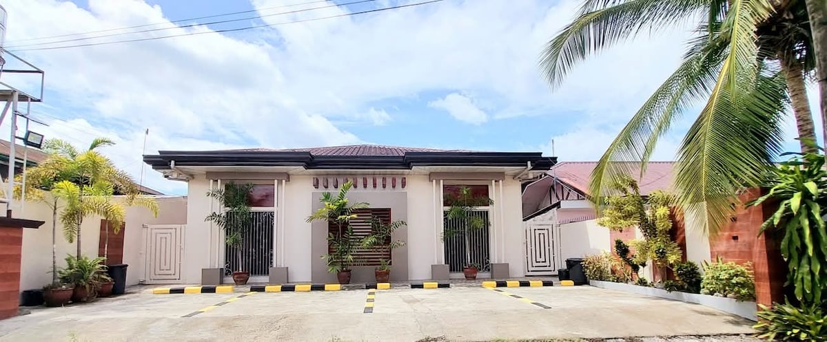 A spacious exterior view of the apartment building is presented, featuring a well-maintained facade with vibrant greenery and potted plants. The structure is flanked by palm trees under a bright sky, and a clear pathway leads to the entrance, providing a welcoming atmosphere.