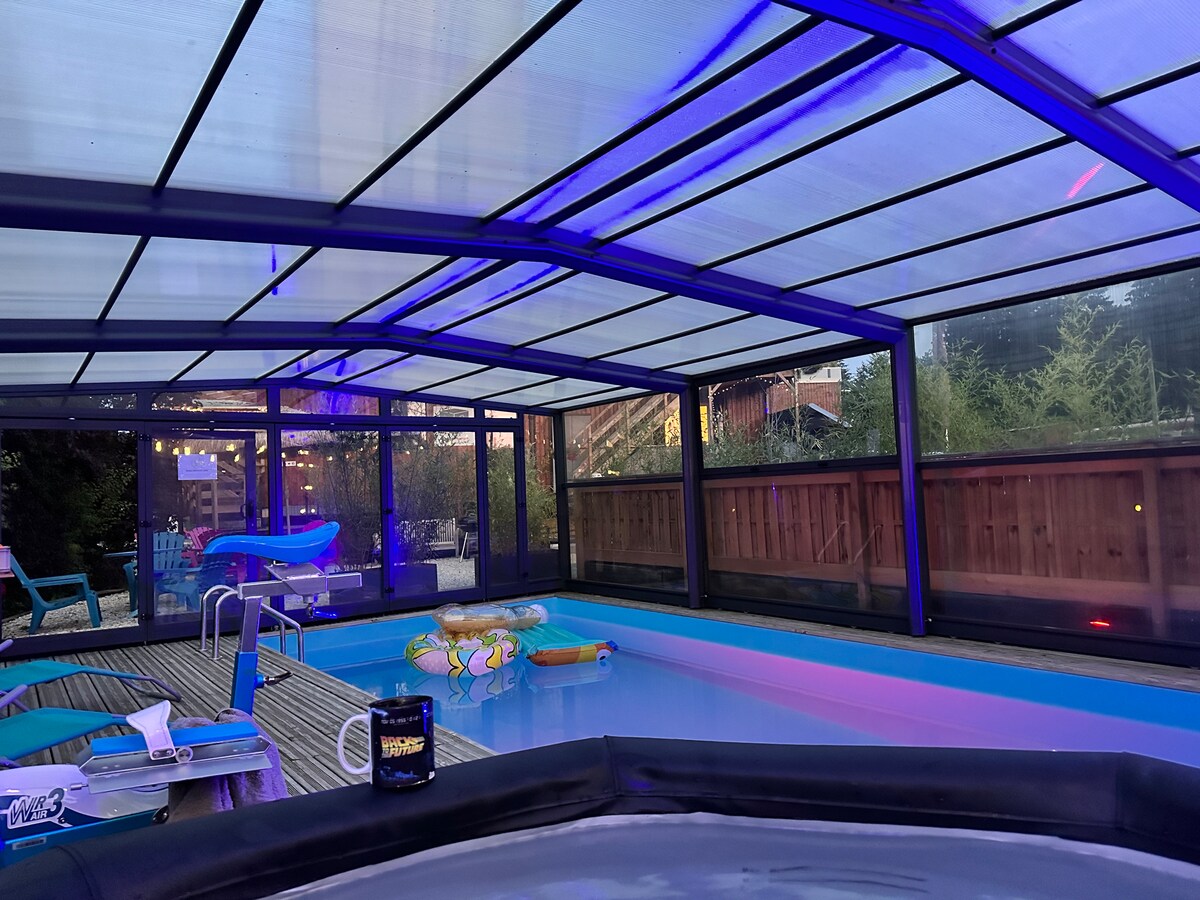 An indoor pool area is enclosed with transparent panels, allowing natural light to fill the space. A jacuzzi is visible in the foreground, surrounded by colorful lounge floats. Soft purple lighting creates a calming ambiance, and chairs are arranged nearby for relaxation.