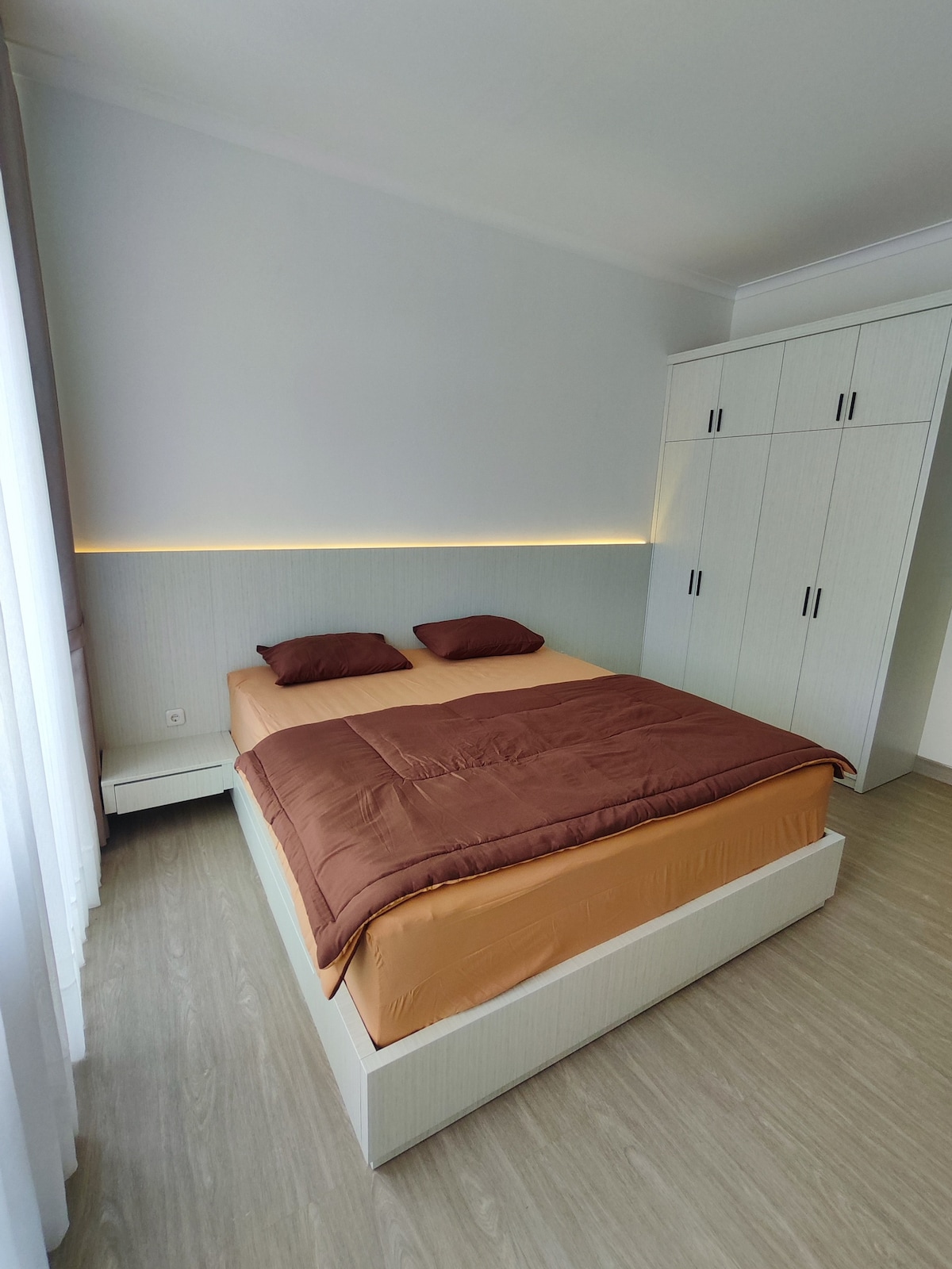 A bright sleeping area is furnished with a spacious bed dressed in warm-toned bedding. Soft lighting is integrated along the wall, while a built-in wardrobe offers storage. Natural light enters through the sheer curtains, complementing the minimalist decor.