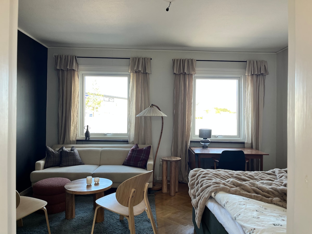 A cozy living space is highlighted by two large windows allowing natural light. A light-colored sofa is combined with wooden stools and a round table, while a desk with a chair occupies one corner. A spacious bed with a soft blanket is visible in the background.