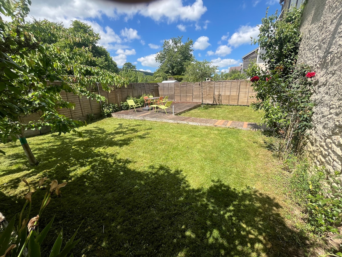 The garden area features a well-maintained lawn surrounded by trees and shrubbery. A wooden terrace is visible, equipped with outdoor furniture for relaxation. Bright chairs are positioned under the sun, providing a pleasant spot for enjoying the outdoors.