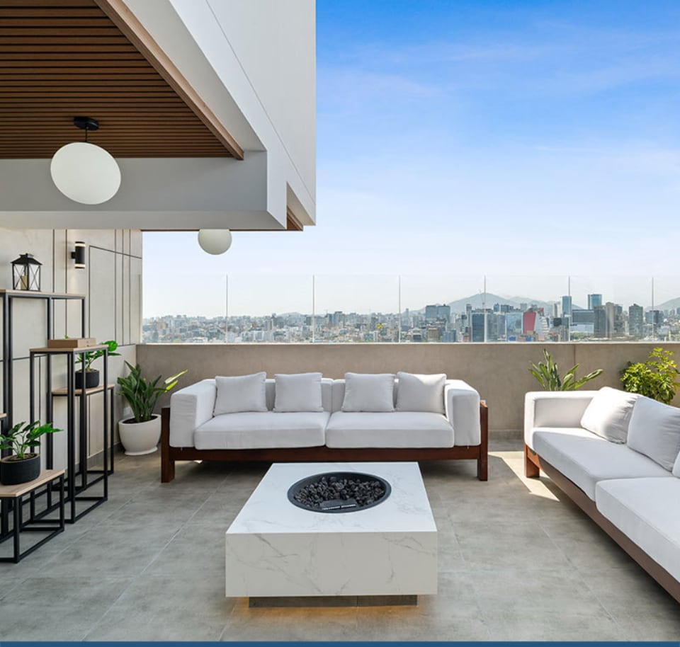 An outdoor terrace features modern furnishings, including a spacious sectional sofa and a coffee table with a fire pit. Green plants are arranged in planters, and views of the city skyline are visible in the background, complemented by a clear blue sky.