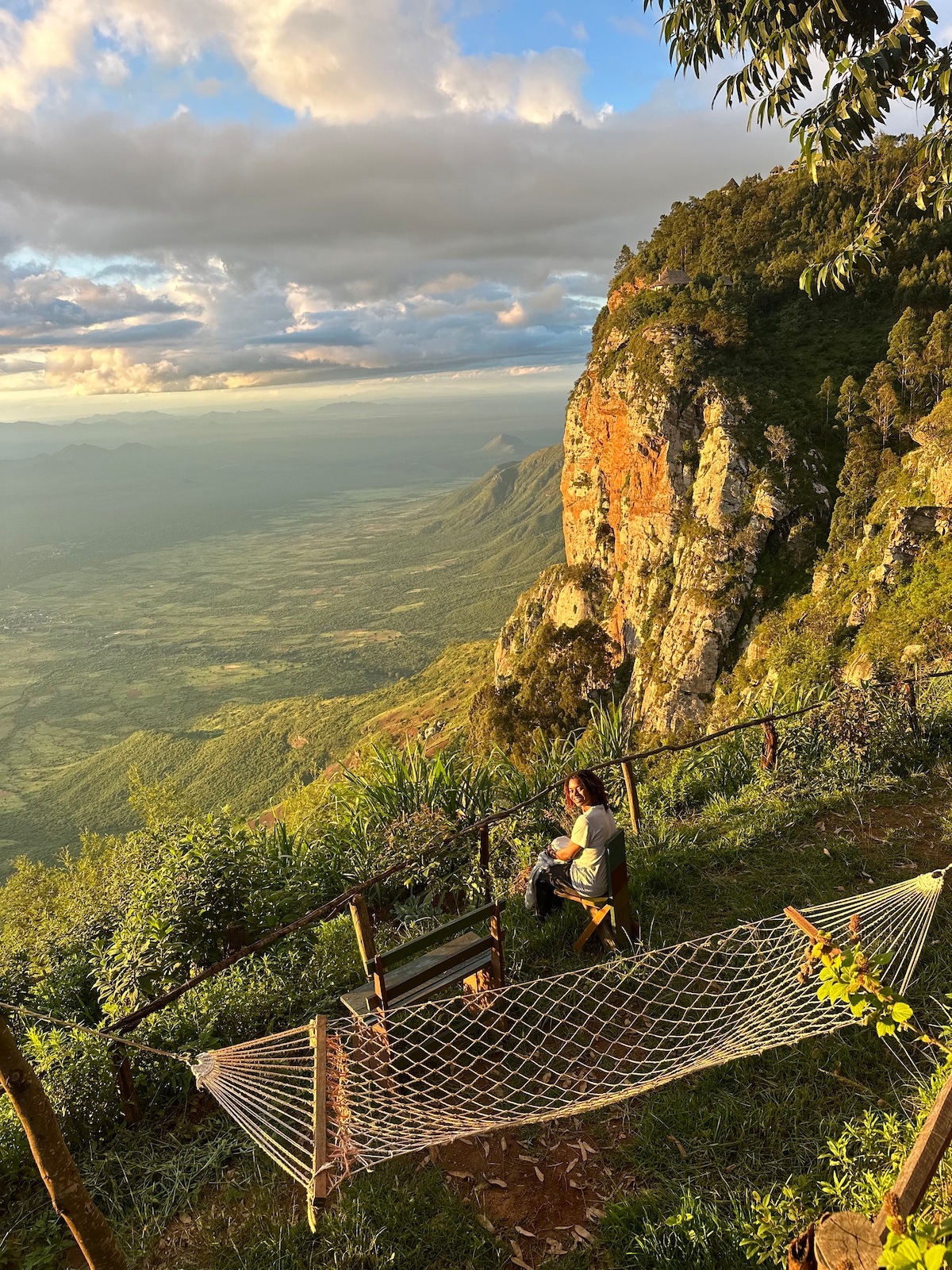 A hammock is positioned on a balcony overlooking a breathtaking landscape of green hills and distant mountains under a partly cloudy sky. The woven texture of the hammock contrasts with the surrounding natural foliage, creating a serene spot for relaxation amidst the stunning views.