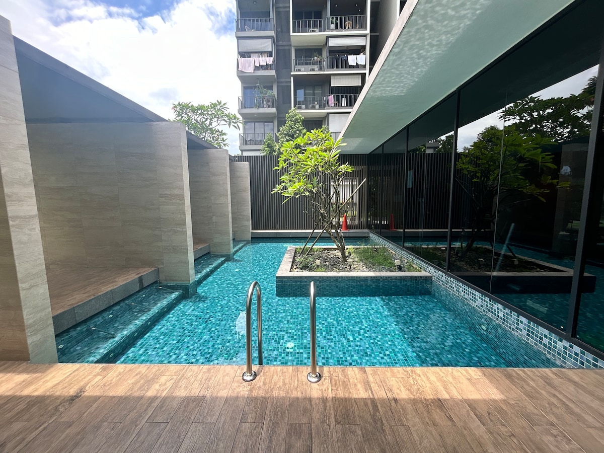 A serene pool area showcases turquoise water, surrounded by a natural stone wall and verdant plants. The peaceful setting is complemented by clear blue skies and modern building facades in the background, creating a calm atmosphere for relaxation.