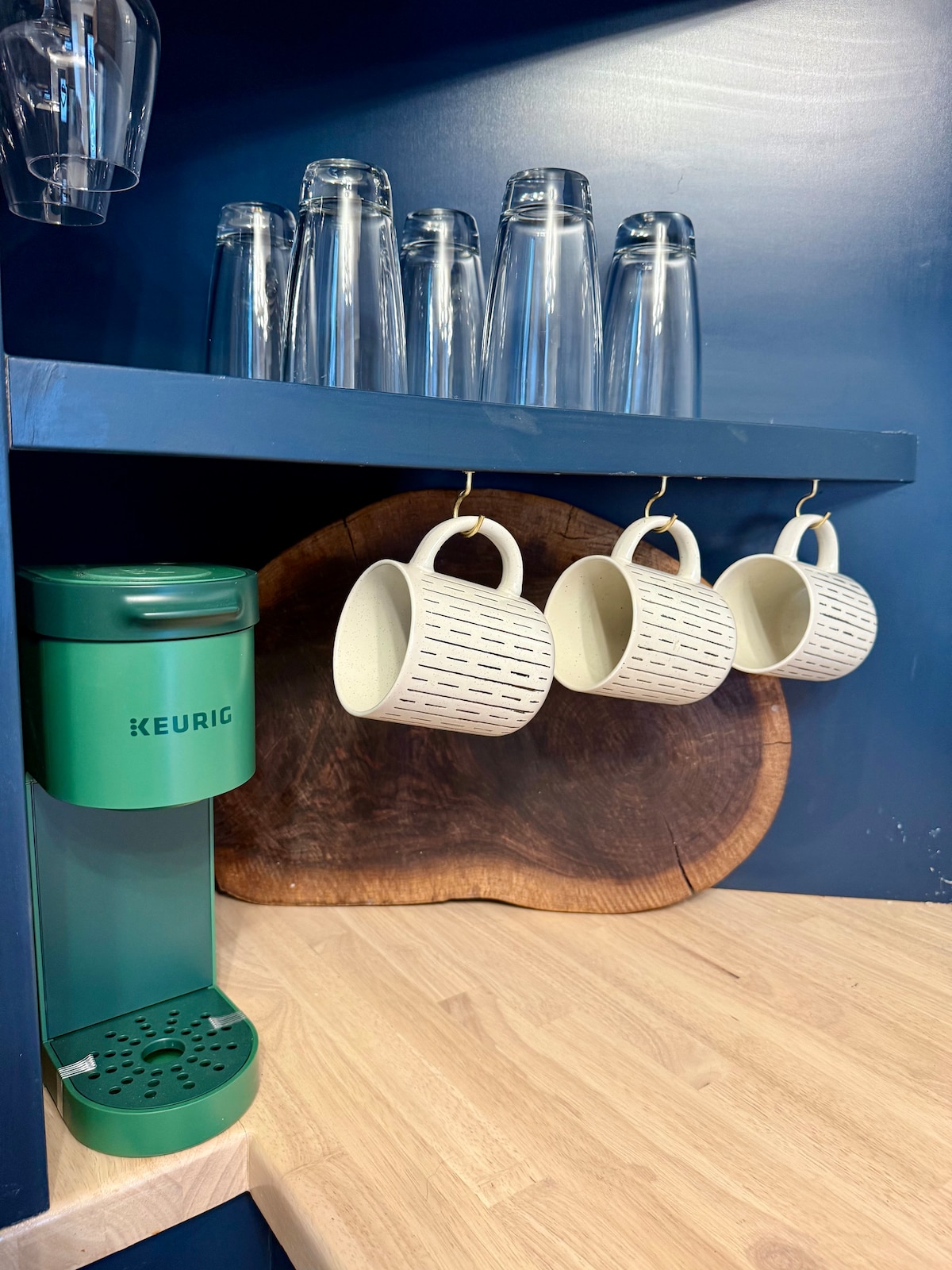 A coffee station is featured with a green Keurig machine positioned next to a wooden cutting board. Three patterned mugs hang from hooks above a light wooden countertop, with several clear glasses arranged on an upper shelf.