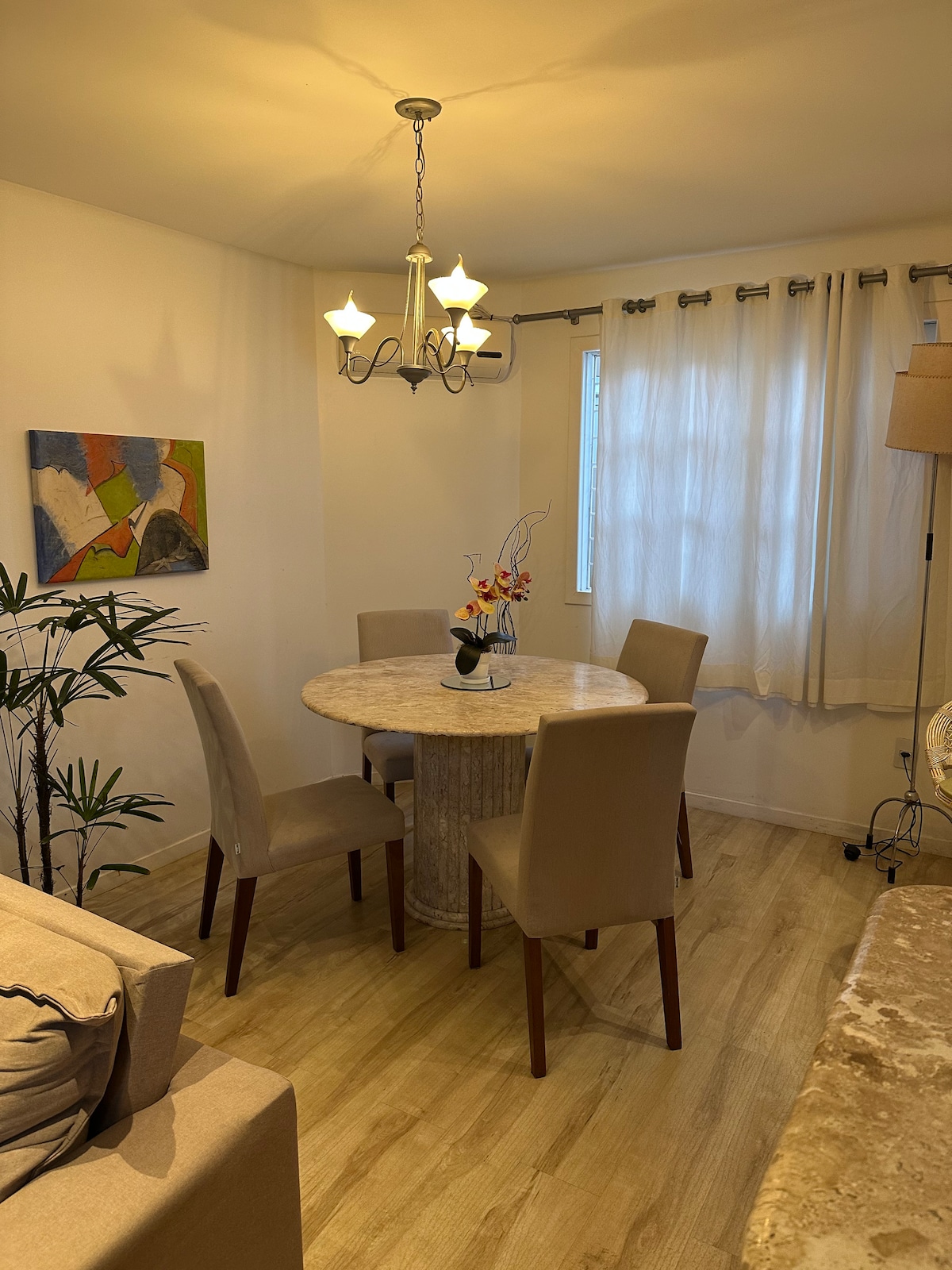 A dining area features a round marble table surrounded by four beige chairs. A chandelier hangs overhead, providing soft lighting. Natural light enters through a window covered by sheer curtains, while a patterned wall art piece adds a touch of color to the neutral surroundings.