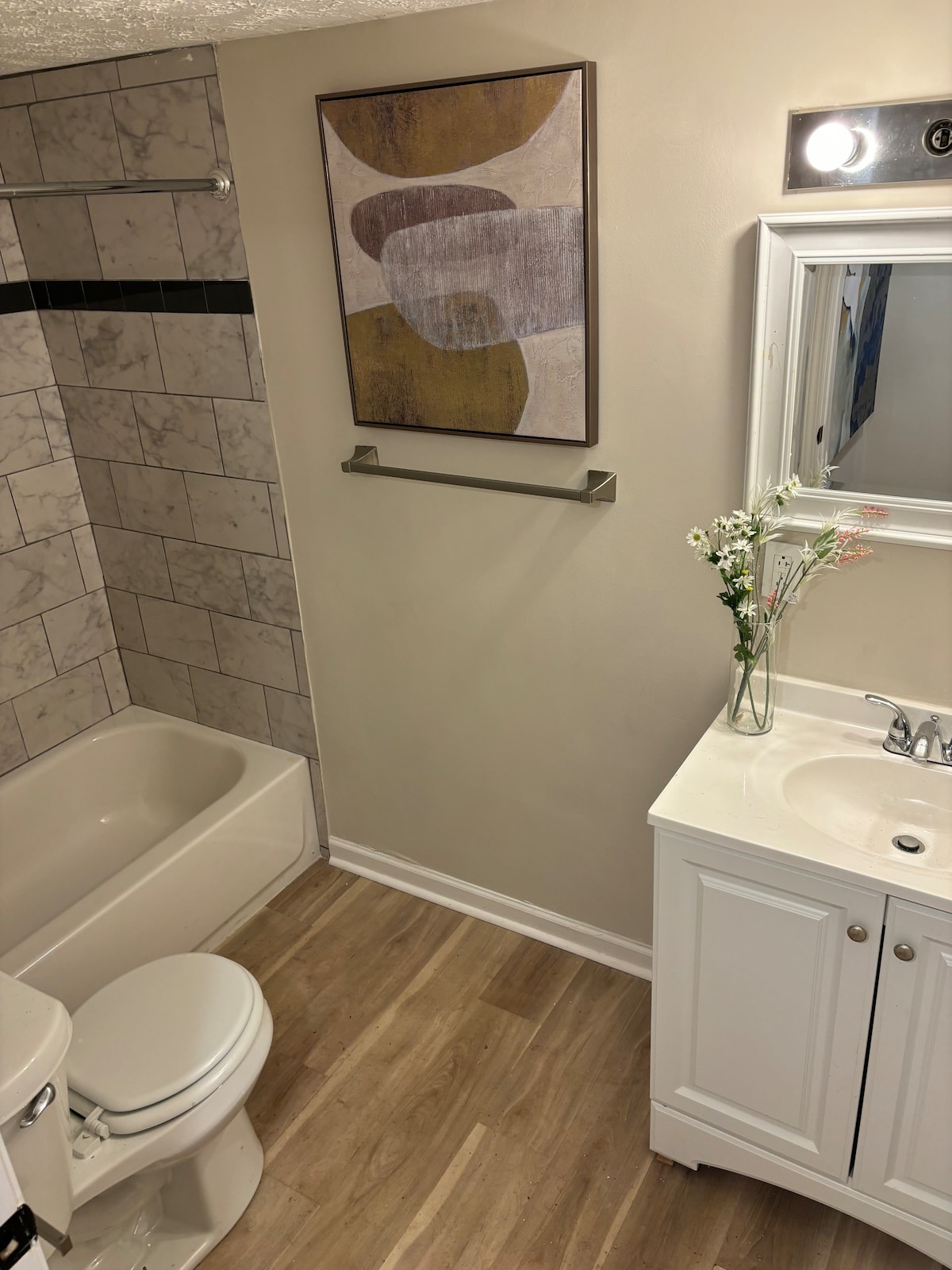 A clean bathroom features a bathtub and a white vanity with a sink. A framed abstract artwork is displayed on the wall above a towel rack. Natural light illuminates the space, highlighting the neutral tones of the tiled walls and floor.