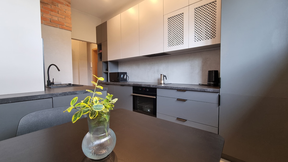 A modern kitchen showcases sleek cabinetry with a combination of light and dark finishes. A countertop extends across the space, complemented by a potted plant. Essential appliances, including an oven and microwave, are visible, with ample storage space offered in cupboards above.