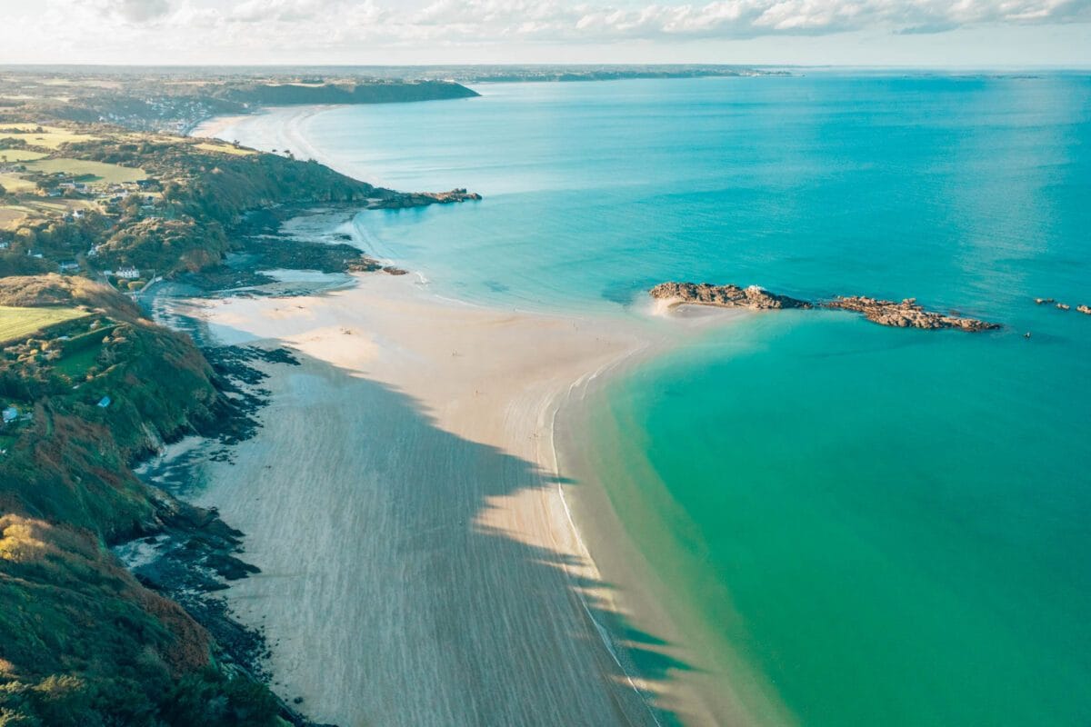 The serene beach landscape stretches along the coastline, with gentle waves lapping at the sandy shore. Lush greenery and cliffs frame the tranquil waters, reflecting shades of blue and green. The horizon meets the sky, dotted with soft clouds, enhancing the coastal scenery.