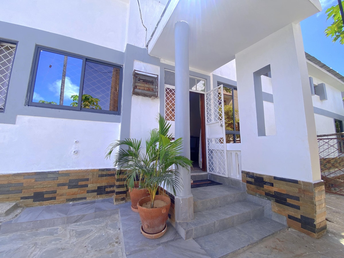 The entrance to the villa is marked by a set of white double doors framed by a small porch. Potted plants are positioned beside the steps, and sunlight reflects off the exterior walls, enhancing the welcoming appearance of the home.