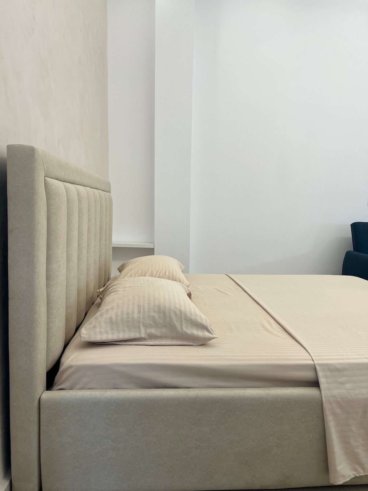 A spacious bed with a soft, upholstered headboard is positioned against a light-colored wall. Neutral-toned bedding is complemented by two neatly arranged pillows. A dark armchair is visible in the background, providing a contrasting element to the serene sleeping space.