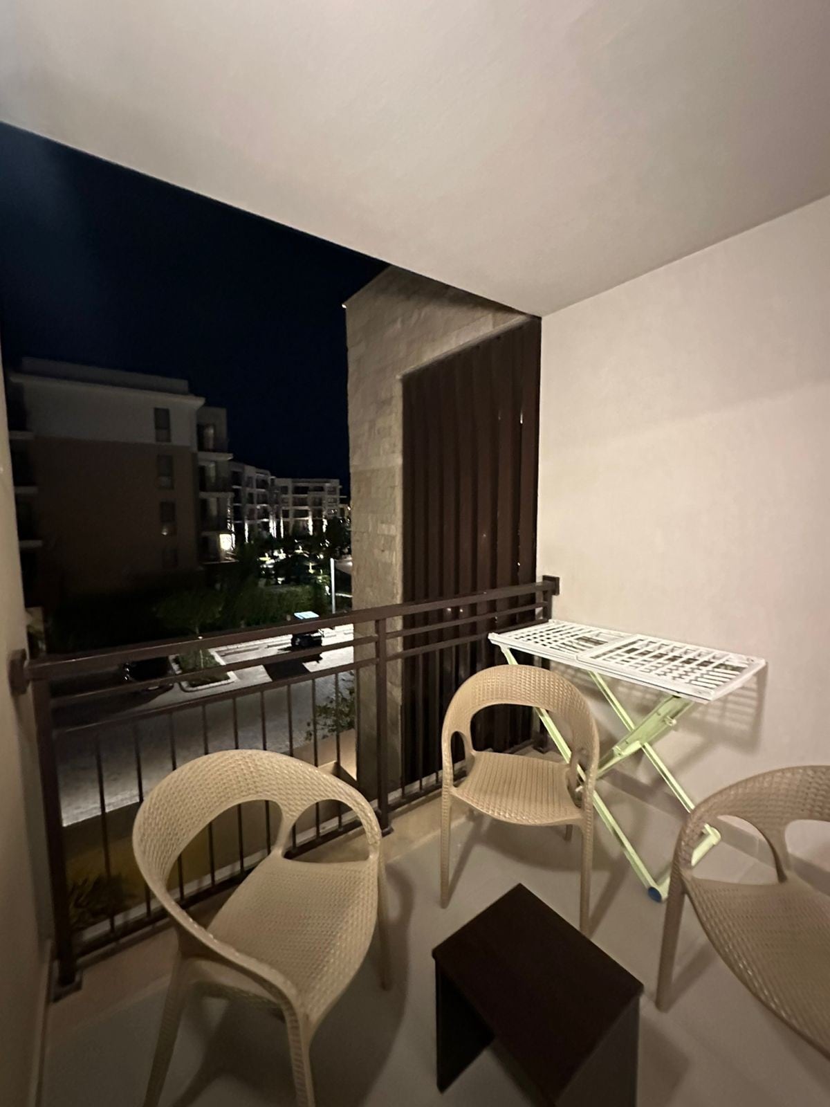 A small balcony is depicted, featuring two light-colored chairs and a compact table. To one side, a drying rack is positioned against the wall. The surrounding area is subtly illuminated, with buildings visible in the background under a night sky.