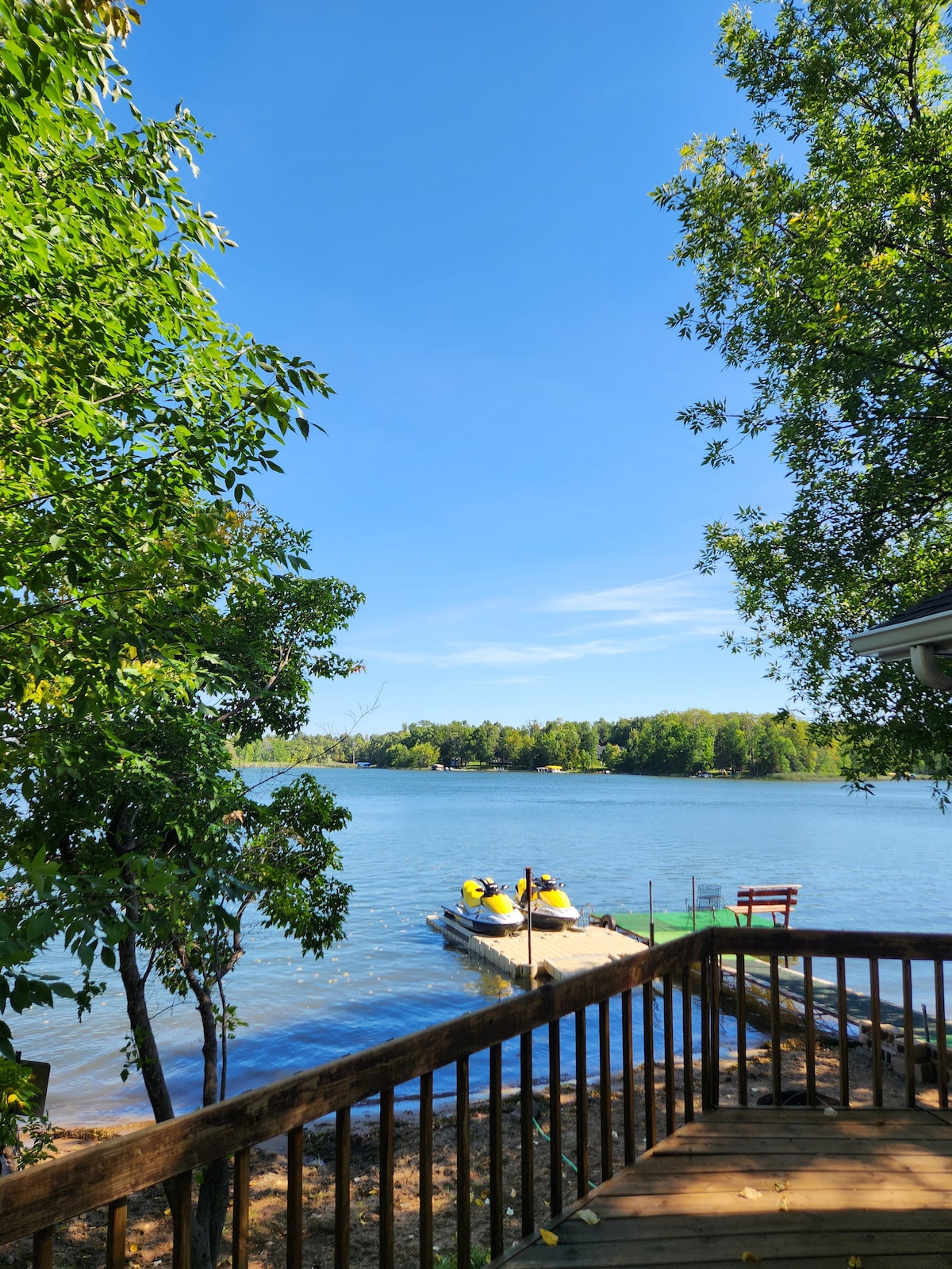 A scenic view of the tranquil lake is framed by lush trees, with a dock visible on the water. Two yellow watercraft are positioned on the dock, reflecting the clear blue sky above and the greenery surrounding the lake.