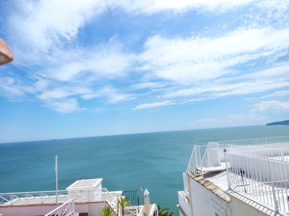A panoramic view of the sea is captured from a terrace, displaying a vast expanse of blue waters meeting a clear sky. The coastline is visible in the distance, while a white railing adorns the terrace, enhancing the overall tranquil atmosphere.