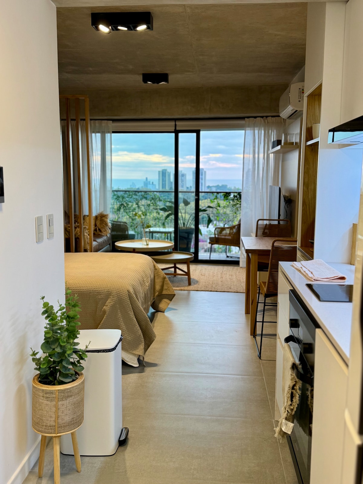 A modern studio apartment is visible, showcasing a spacious bed and a cozy seating area. Large glass doors provide a view of the city skyline and open onto a balcony. A minimalist kitchen is neatly arranged with light-colored cabinetry and essential appliances.