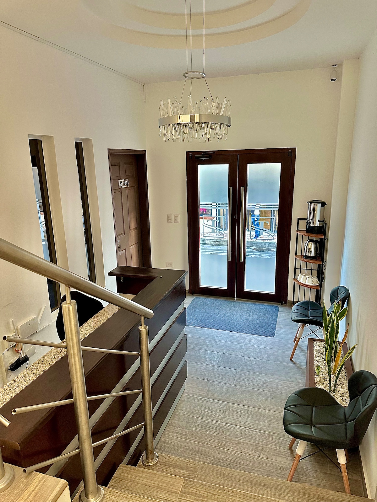 A welcoming entrance area features a contemporary reception desk and two stylish black chairs, positioned beside a small shelf with a coffee station. Large glass doors offer access to the outside, allowing natural light to fill the space.