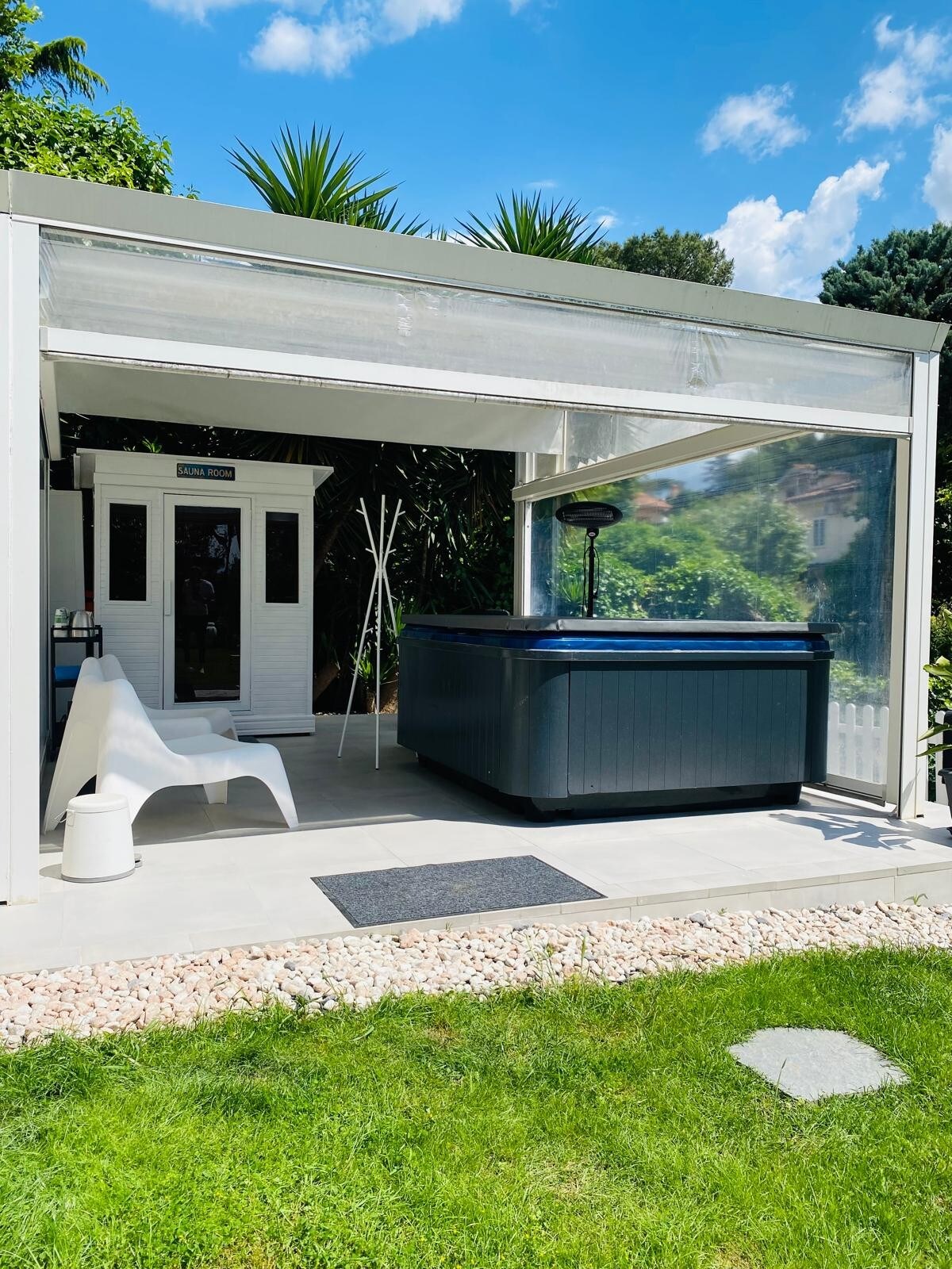 A spa area is showcased with a Jacuzzi hot tub situated beneath an awning, providing shade. Nearby, a white chair sits on the patio beside a small table, with a lush green garden in the background and a clear blue sky overhead.