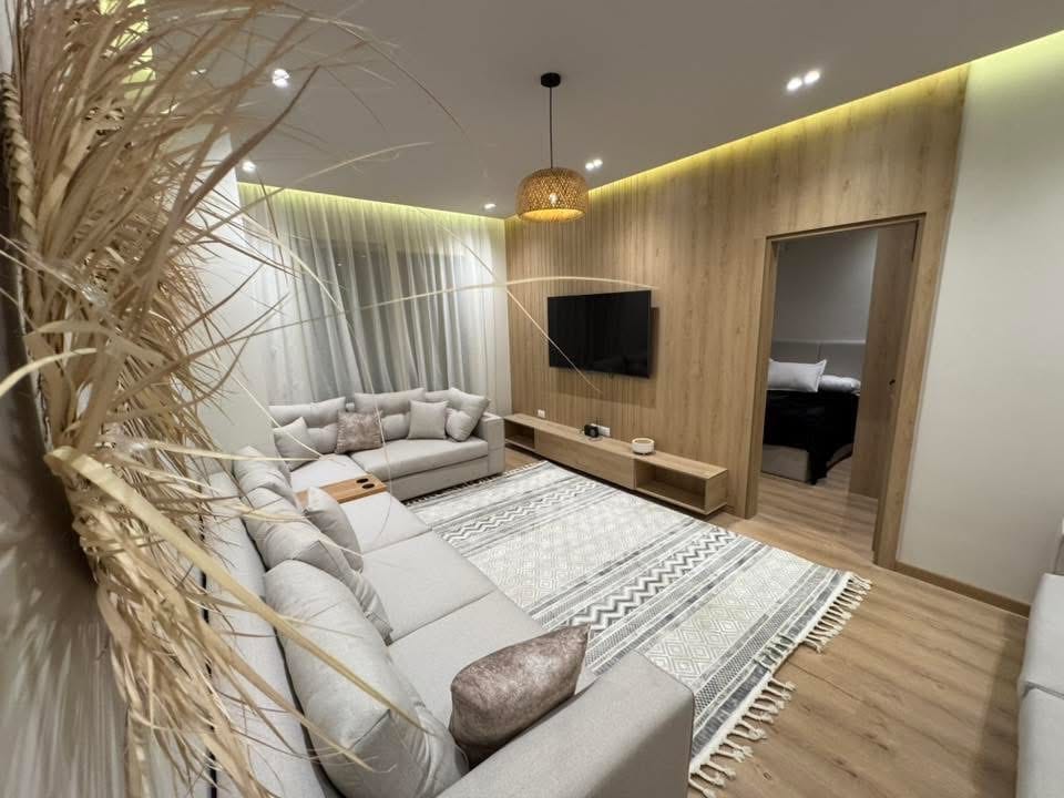 A modern living area features a spacious sectional sofa and a soft area rug. A wall-mounted television is positioned above a light wooden media console. Natural light filters in through sheer curtains, contributing to a relaxed ambiance, while a doorway leads to an adjacent room.