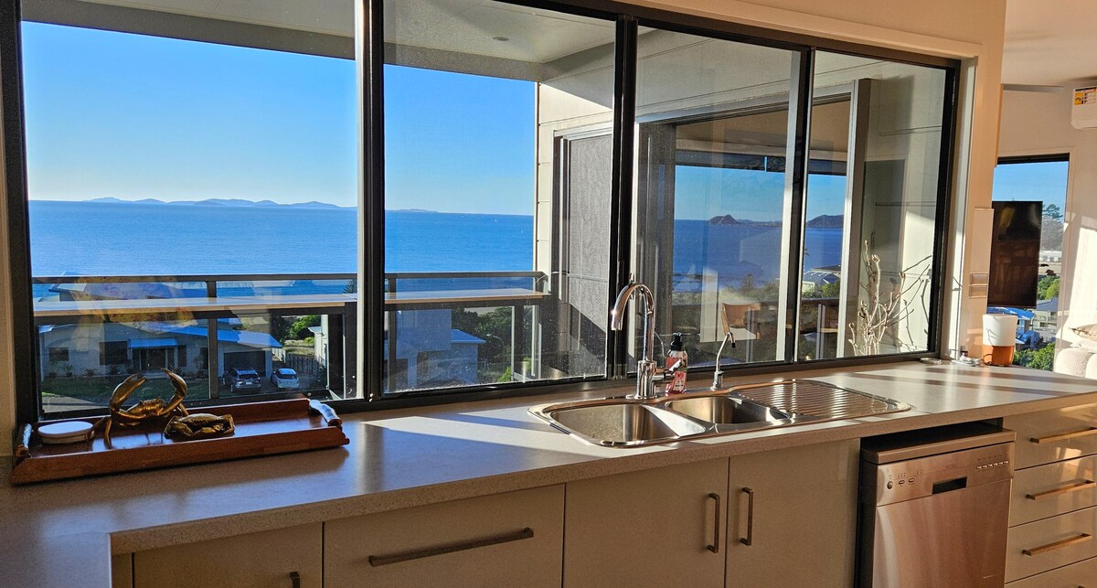 A modern kitchen is displayed with expansive windows providing stunning views of the ocean. Light streams in, illuminating the sleek countertops and stainless steel appliances. The sink area includes a double basin, while a decorative tray is placed nearby.