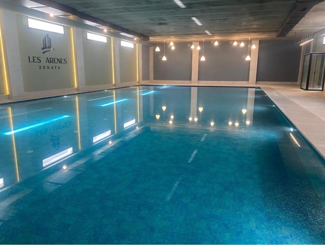 An indoor swimming pool is presented, featuring a large, clear water surface that reflects ceiling lights. The pool area is surrounded by smooth tiles, with a modern design emphasized by minimalist lighting and soft hues on the walls.