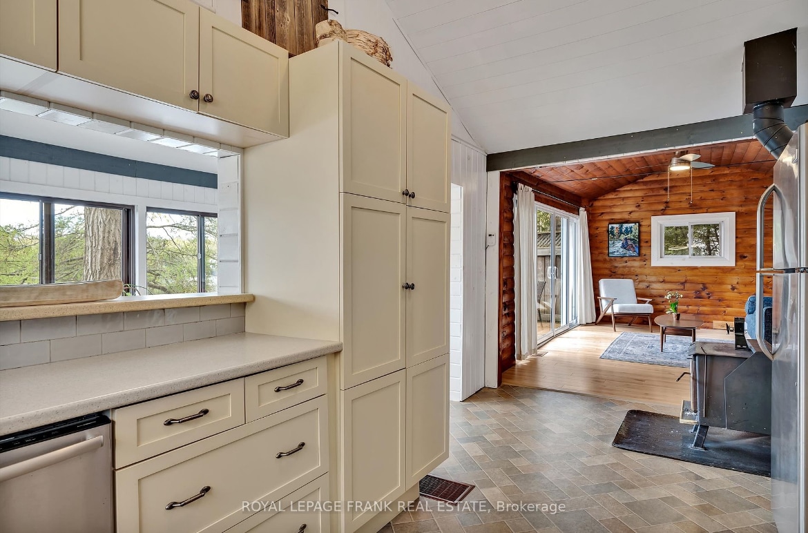 The image showcases a kitchen area with light-colored cabinetry and a textured countertop. A large window allows natural light to fill the space, with an adjacent room visible that features wooden walls and a cozy seating arrangement.