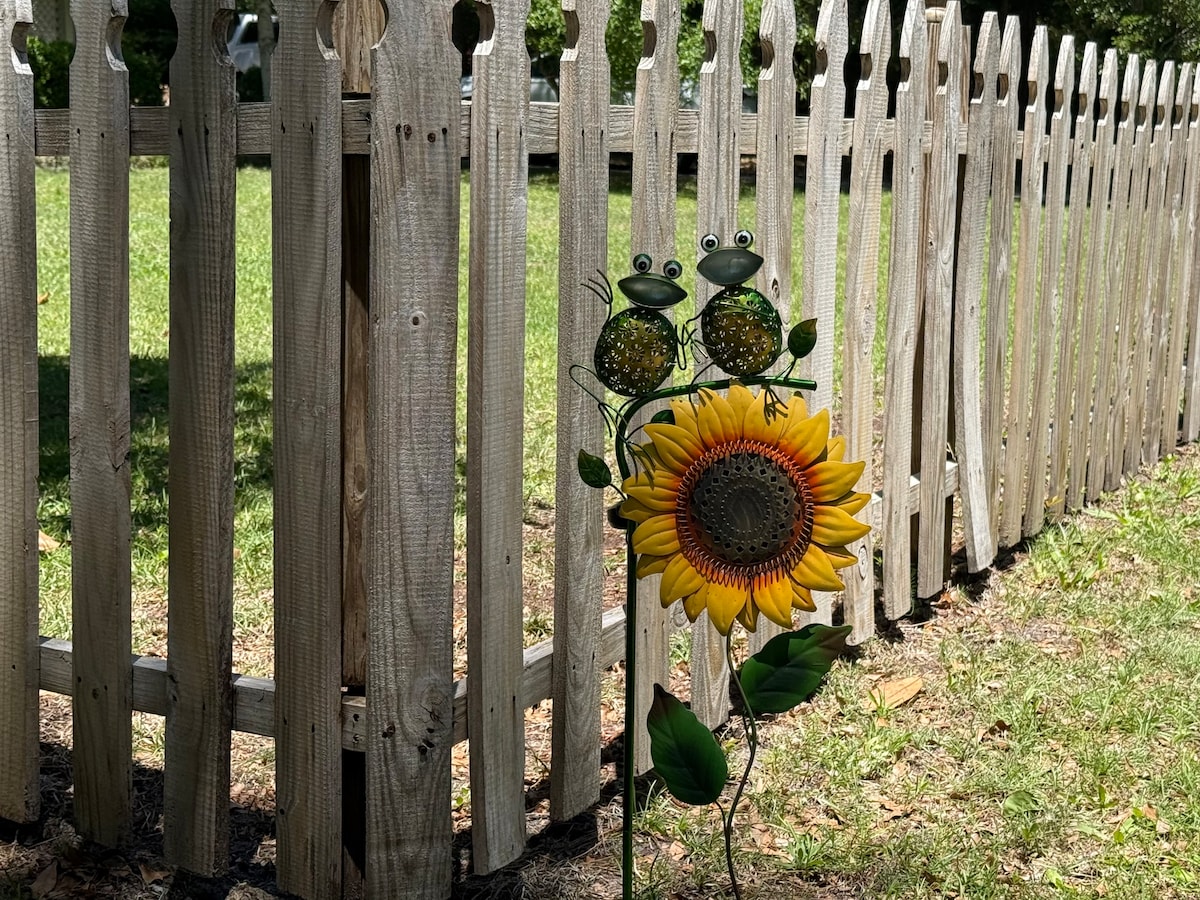 A charming decorative piece is displayed in front of a wooden picket fence. Two frog figurines adorn a vibrant sunflower, adding a playful touch to the greenery. The garden area features a manicured lawn, creating a welcoming atmosphere in the outdoor space.