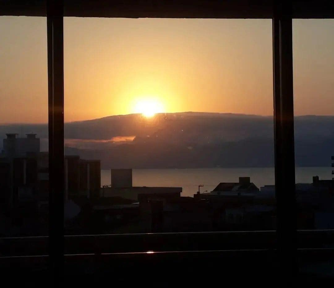 The image captures a sunrise viewed from a window, with a golden orb peeking over a horizon of clouds. Buildings in the foreground create a silhouette against the gentle hues of orange and yellow reflected on the sea.