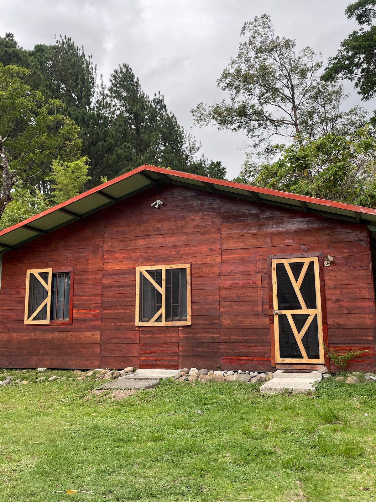 A wooden cabin with a deep red finish is set against a backdrop of greenery. It features two windows and a door, all framed with light wood. The floor is paved with stone steps leading to the entrance. Surrounding vegetation adds to the natural ambiance.
