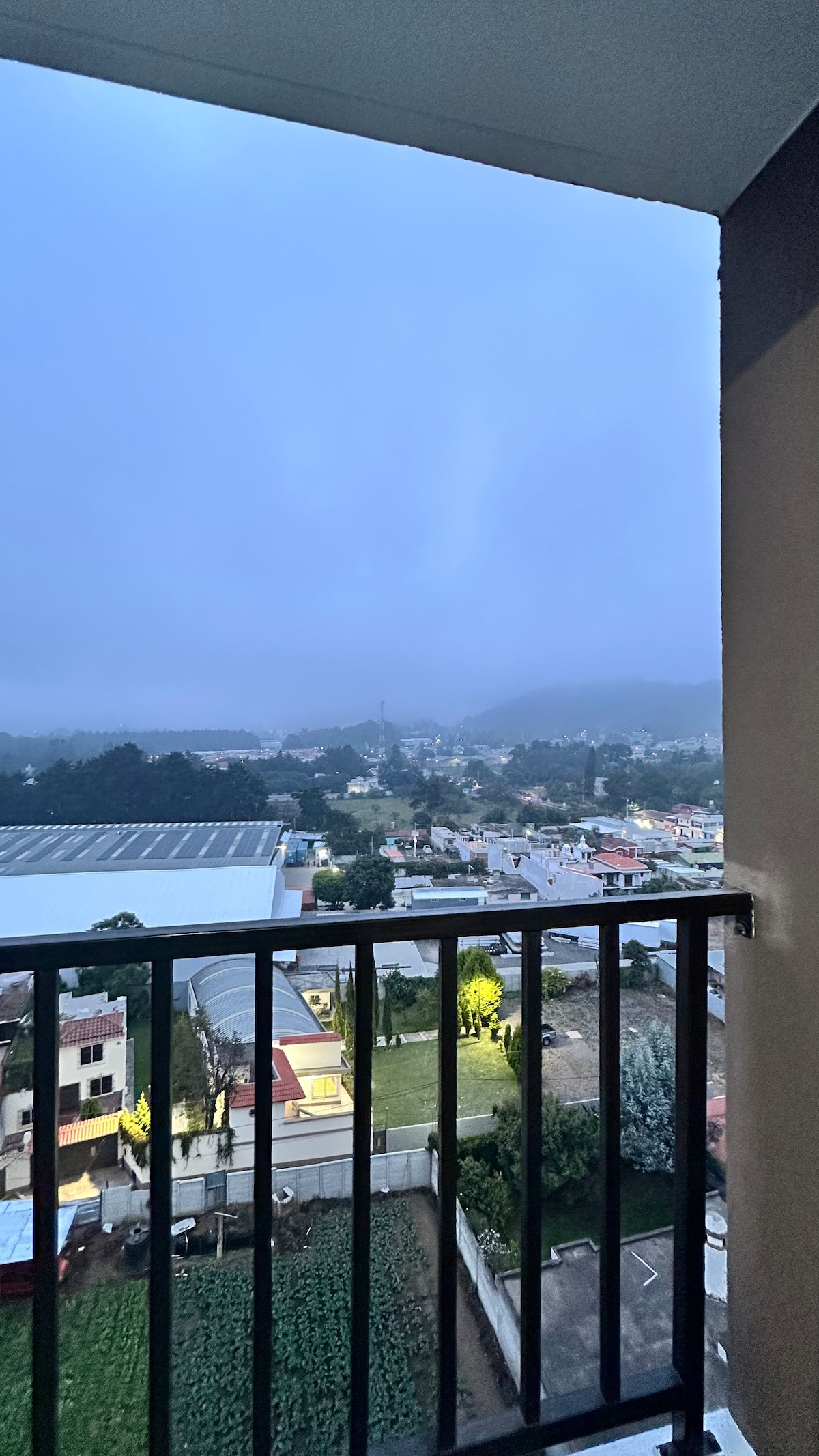 A scenic view from a balcony captures a misty morning landscape. The image features a distant town surrounded by rolling hills and greenery, with rooftops peeking through the fog. A black railing frames the view, enhancing the perspective of the surroundings.
