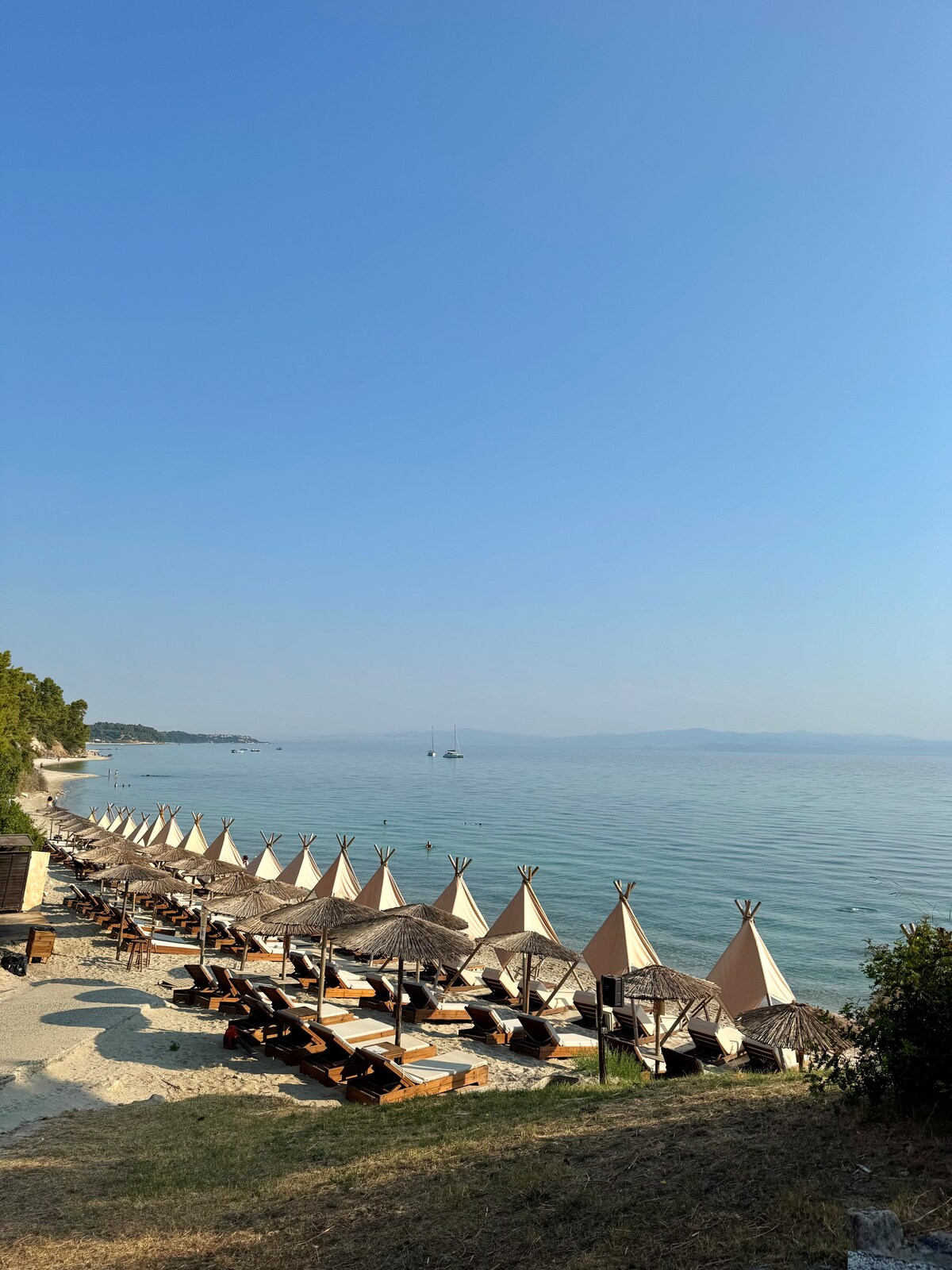 A serene coastal view showcases a row of sunbeds beneath elegant umbrellas along a sandy beach. The calm sea glistens under a clear blue sky, with a distant sailboat visible on the horizon, creating a peaceful and inviting atmosphere.