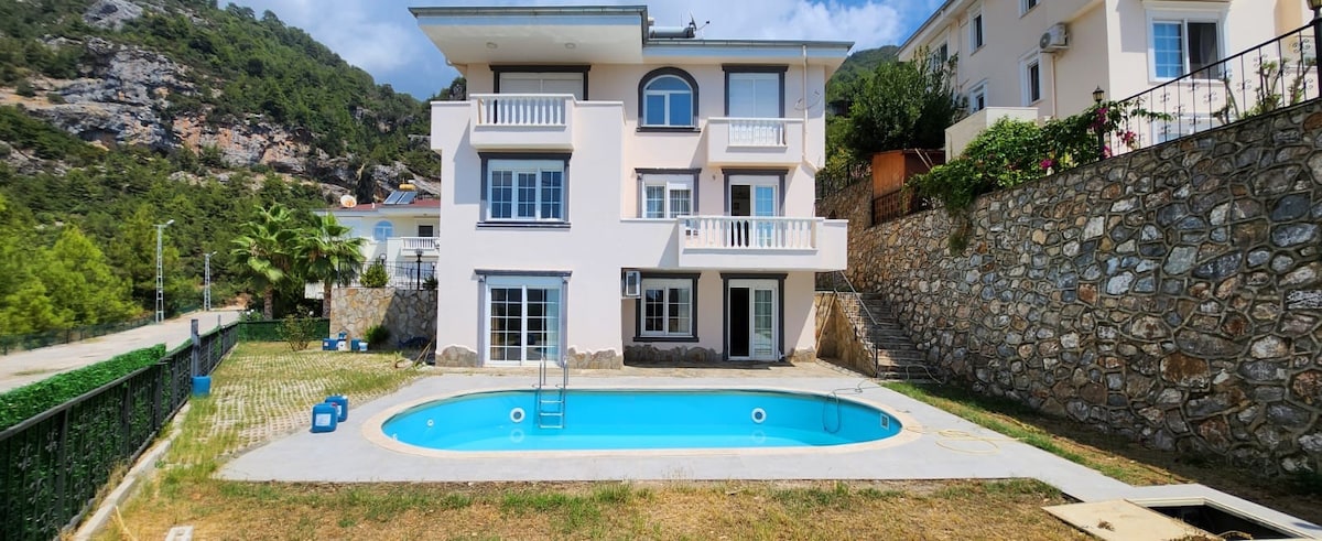 A spacious villa stands with an inviting swimming pool in front. The structure features multiple balconies and large windows, allowing natural light to fill the interior. Surrounding greenery and a stone wall create a serene outdoor environment, offering a connection to nature.