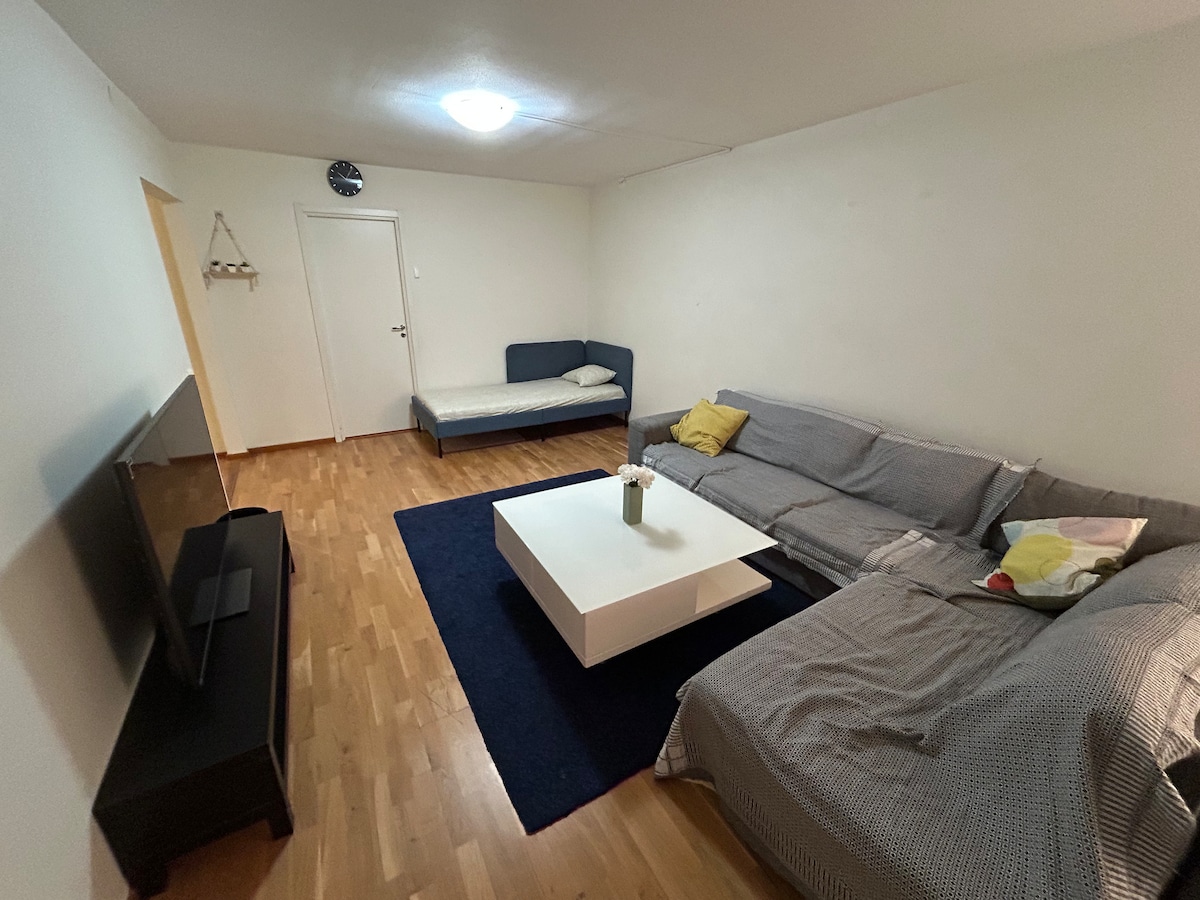 A spacious living area features light-colored wooden flooring and neutral walls. A large sectional sofa is paired with a small couch. A white coffee table sits at the center, complemented by a decorative vase. A television is positioned on a dark cabinet against one wall.
