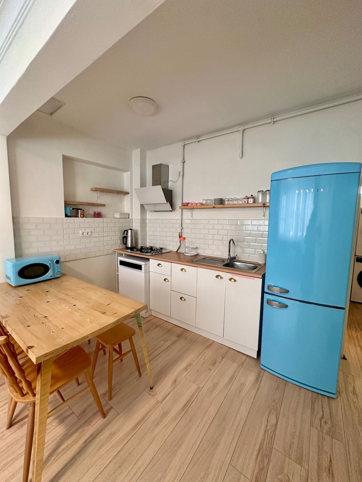 A compact kitchen area features a bright blue refrigerator and modern appliances. A wooden dining table with three chairs provides seating, while white cabinetry and light-colored tiles enhance the space. Natural light filters in, creating an inviting environment for meal preparation and dining.