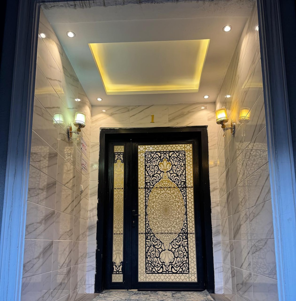 An elegantly designed entrance is showcased, featuring a decorative black door adorned with intricate gold patterns. The walls are finished in light-colored tiles, complemented by soft, recessed lighting and wall sconces, creating a welcoming atmosphere. The flooring is made of stylish stone tiles.