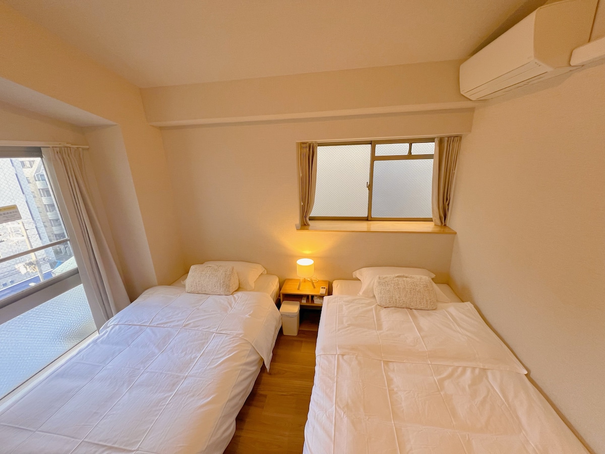 The room features two spacious beds with crisp white linens, positioned next to a bedside table with a soft lamp. Natural light enters through a window, which is partially draped for privacy, illuminating the neutral-colored walls.