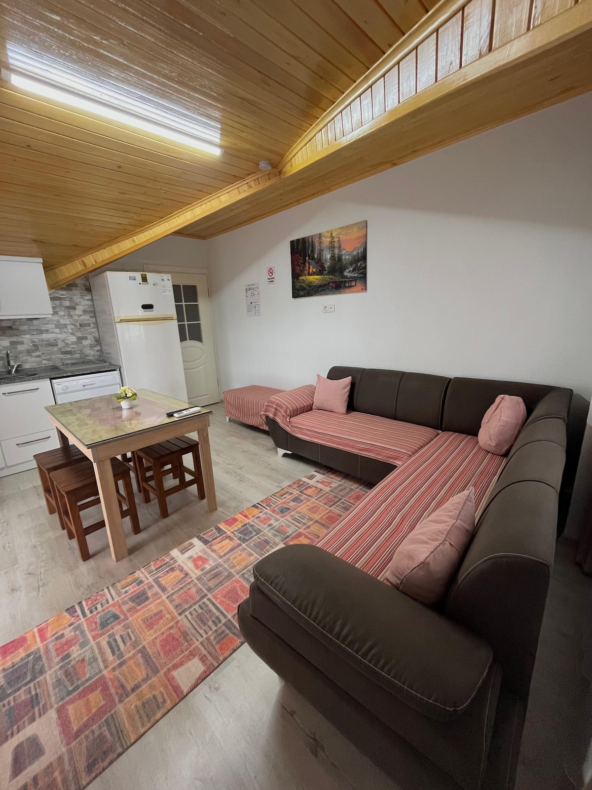 A cozy living space is depicted, featuring a brown sectional sofa with decorative pillows and a small dining table with four wooden chairs. A colorful area rug lies on the floor, while a framed landscape art piece adorns the wall. Natural light enters through the window.