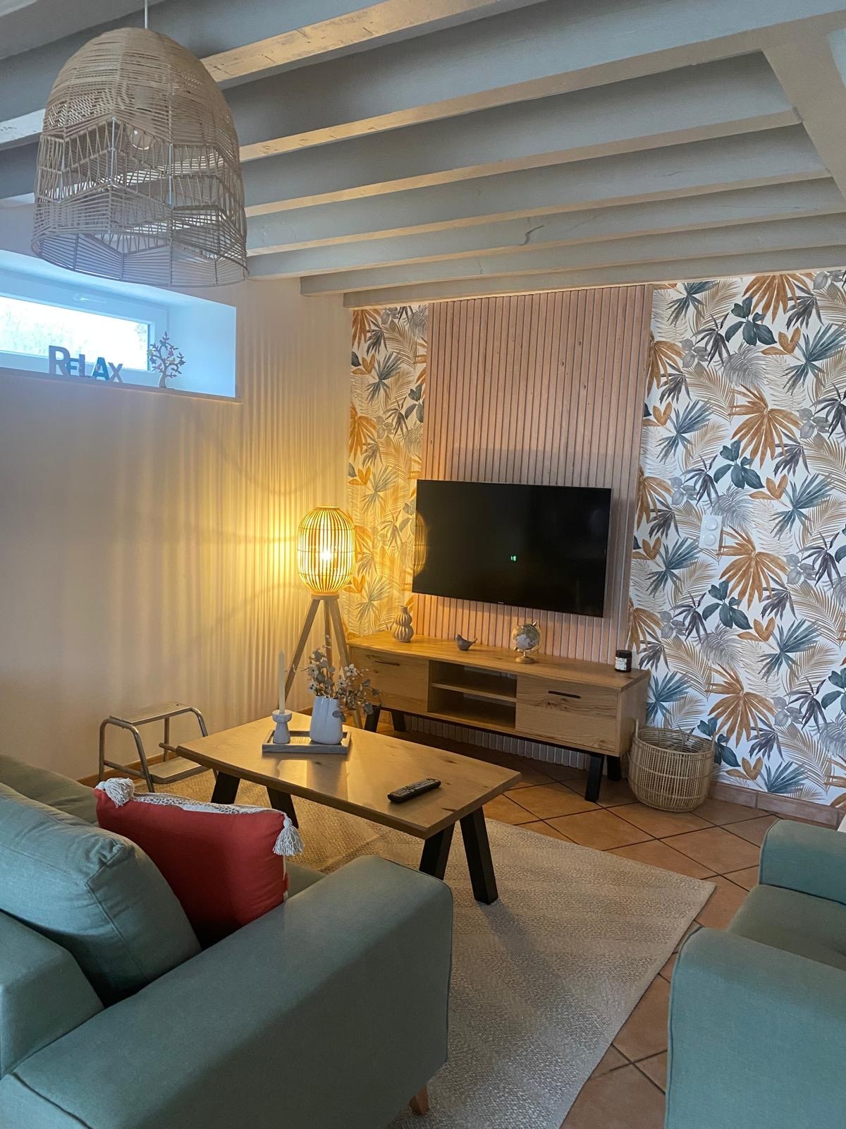 A comfortable seating area is showcased, featuring a soft green sofa with vibrant cushions. A light fixture adds warmth, while a television is mounted against a backdrop adorned with floral wallpaper. Natural light enters through a small window, enhancing the cozy atmosphere.