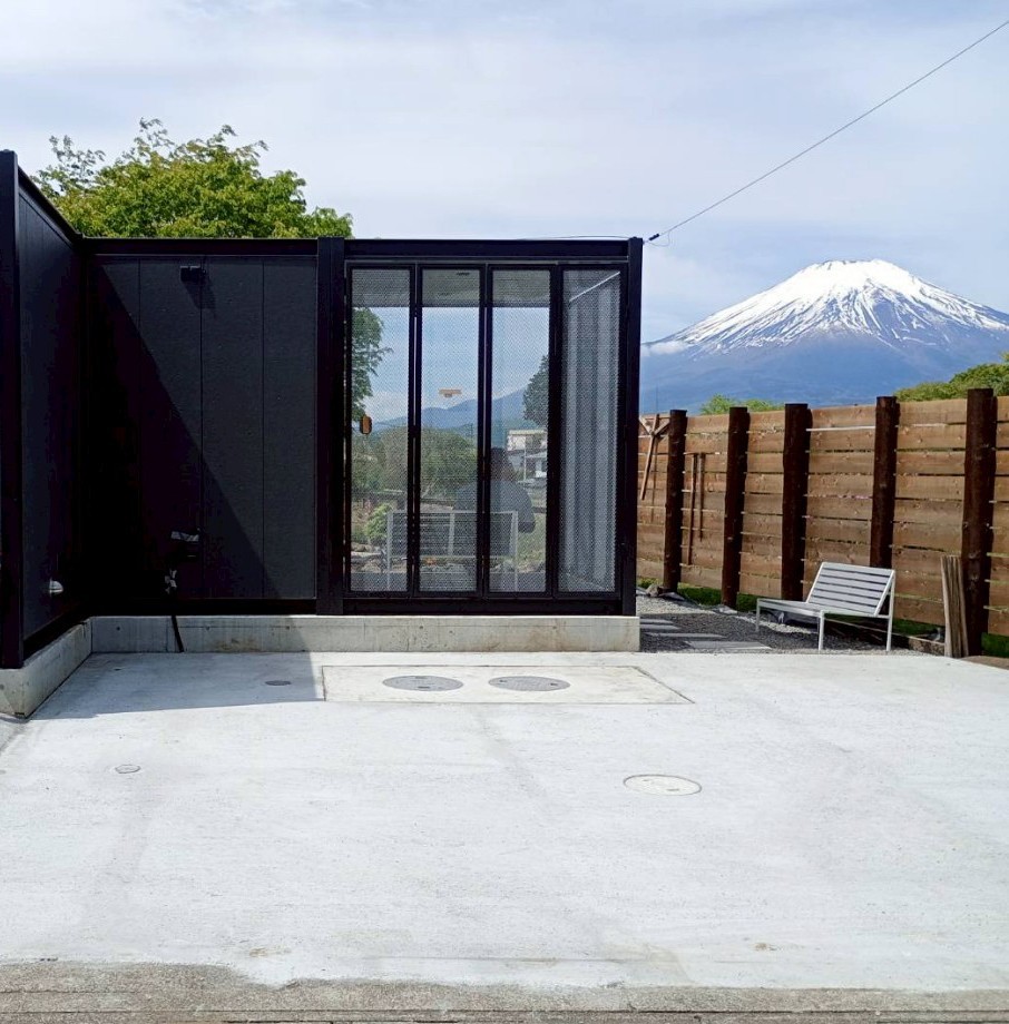 An outdoor area is depicted, featuring a sleek black structure with large glass panels. A view of Mount Fuji rises majestically in the background. A single lounge chair rests on the concrete surface, surrounded by a wooden fence for privacy.