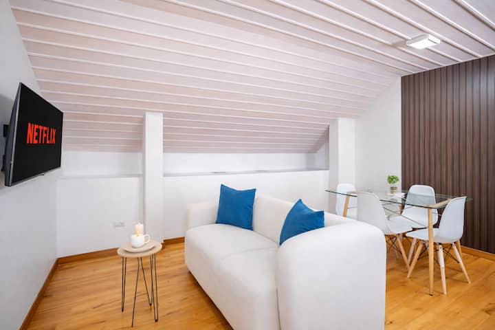 Área social acogedora en ático, con sofá cómodo, comedor moderno y TV con Netflix. Piso de madera y techo inclinado que aporta calidez. Ideal para relajarse o compartir en un ambiente tranquilo.