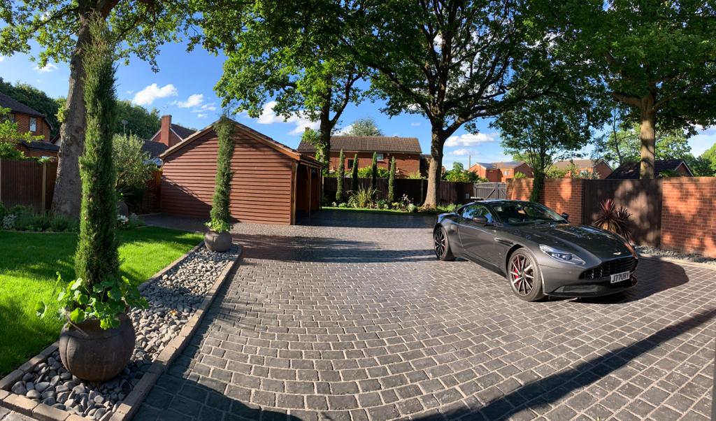 The image features a spacious driveway made of patterned cobblestones, flanked by well-maintained landscaping. A sleek dark car is parked on the right, while a timber-framed carriage house stands in the background, surrounded by lush greenery and clear blue skies.