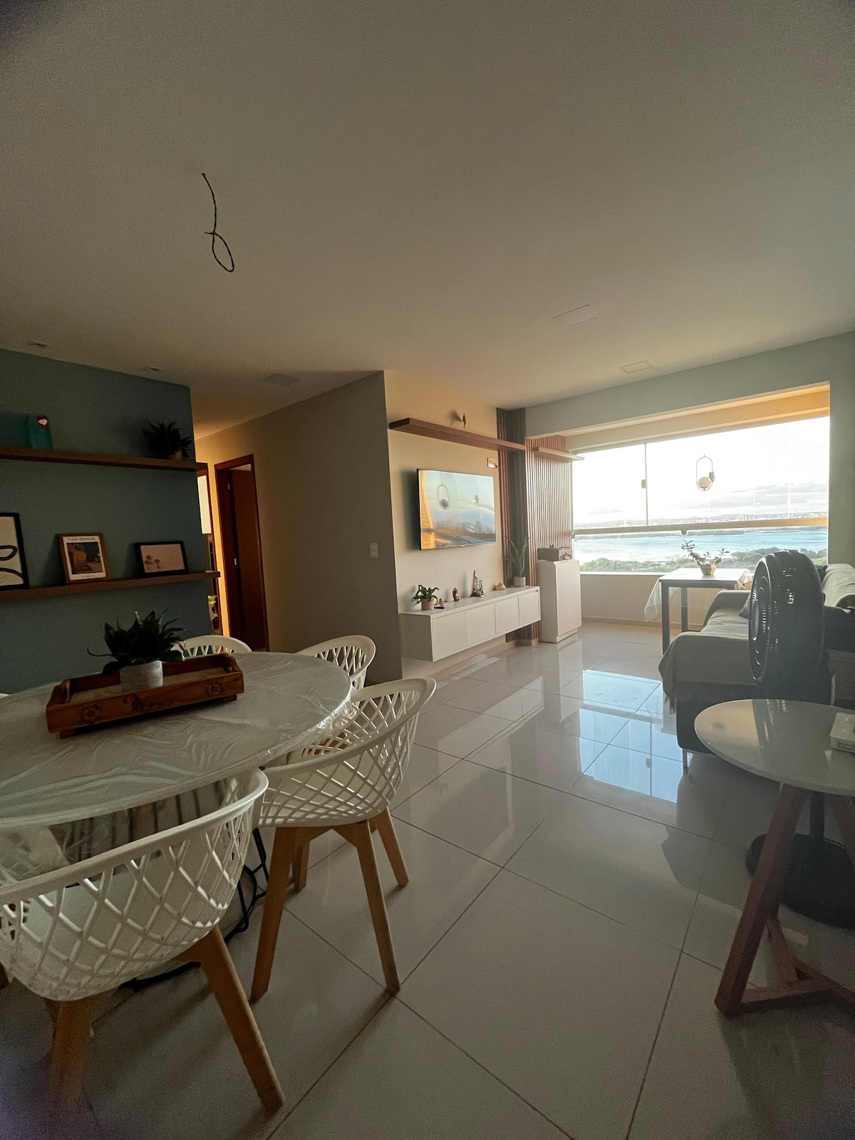 A bright living space features a dining area with white, intricately designed chairs surrounding a round table. A large window allows natural light to fill the room, revealing views of the outside landscape. A comfortable seating area with a sofa and decorative elements is visible across the space.
