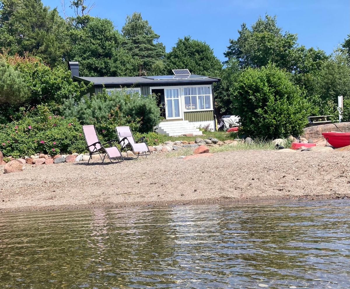 The image captures a private sand beach bordered by lush greenery. Two lounge chairs are positioned on the sandy shore, facing a building with large windows. The clear water reflects the sky, enhancing the serene atmosphere of the location.