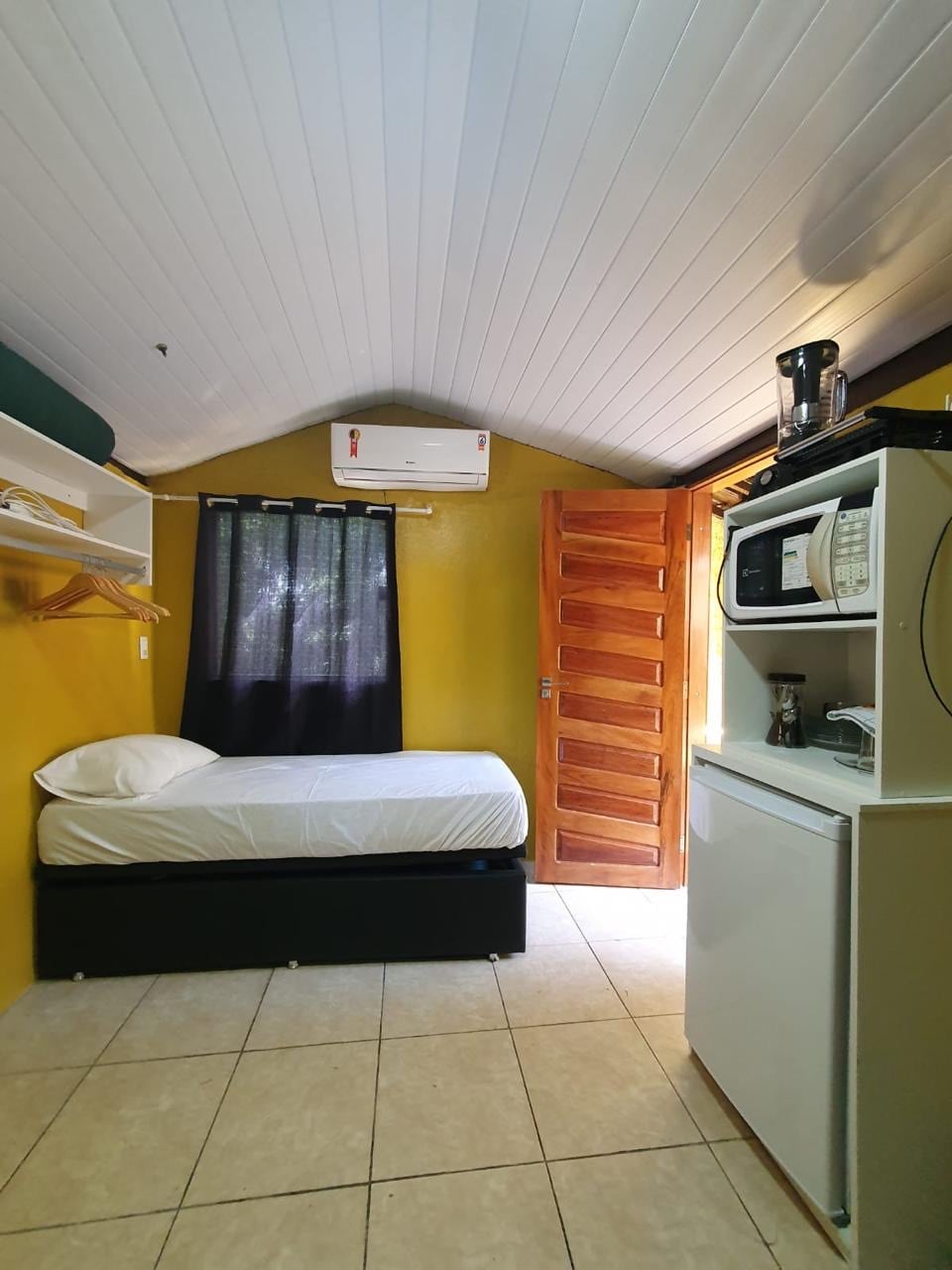 A compact sleeping area features a single bed with white bedding against a vibrant yellow wall. A wooden closet provides hanging space. A small kitchenette, including a microwave and coffee maker, is positioned beside the bed. Natural light enters through a window covered by dark curtains.