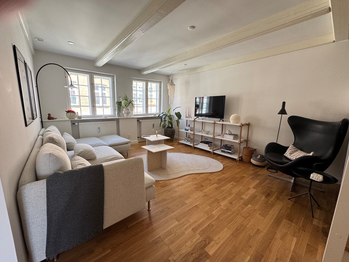 A spacious living area is highlighted by light wood flooring and soft neutral-colored furnishings. A large sectional sofa and a cozy armchair face a modern television stand. Natural light enters through multiple windows, complemented by indoor plants and a round area rug on the floor.