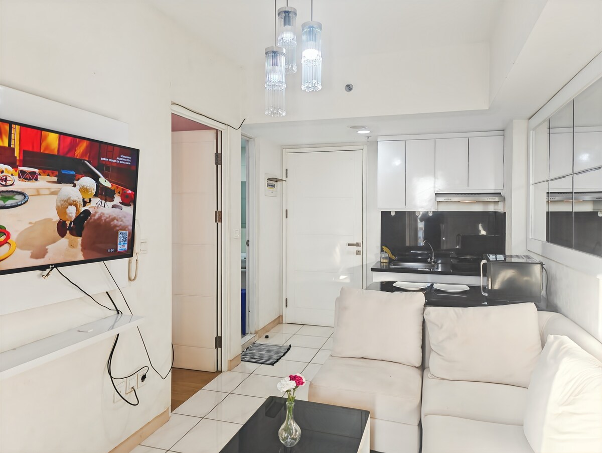 A comfortable living area features a light-colored couch paired with a dark coffee table. A television is mounted on the wall, displaying content. The kitchen is visible in the background, showcasing modern cabinetry and appliances, while a small vase with a flower adds a touch of decor.