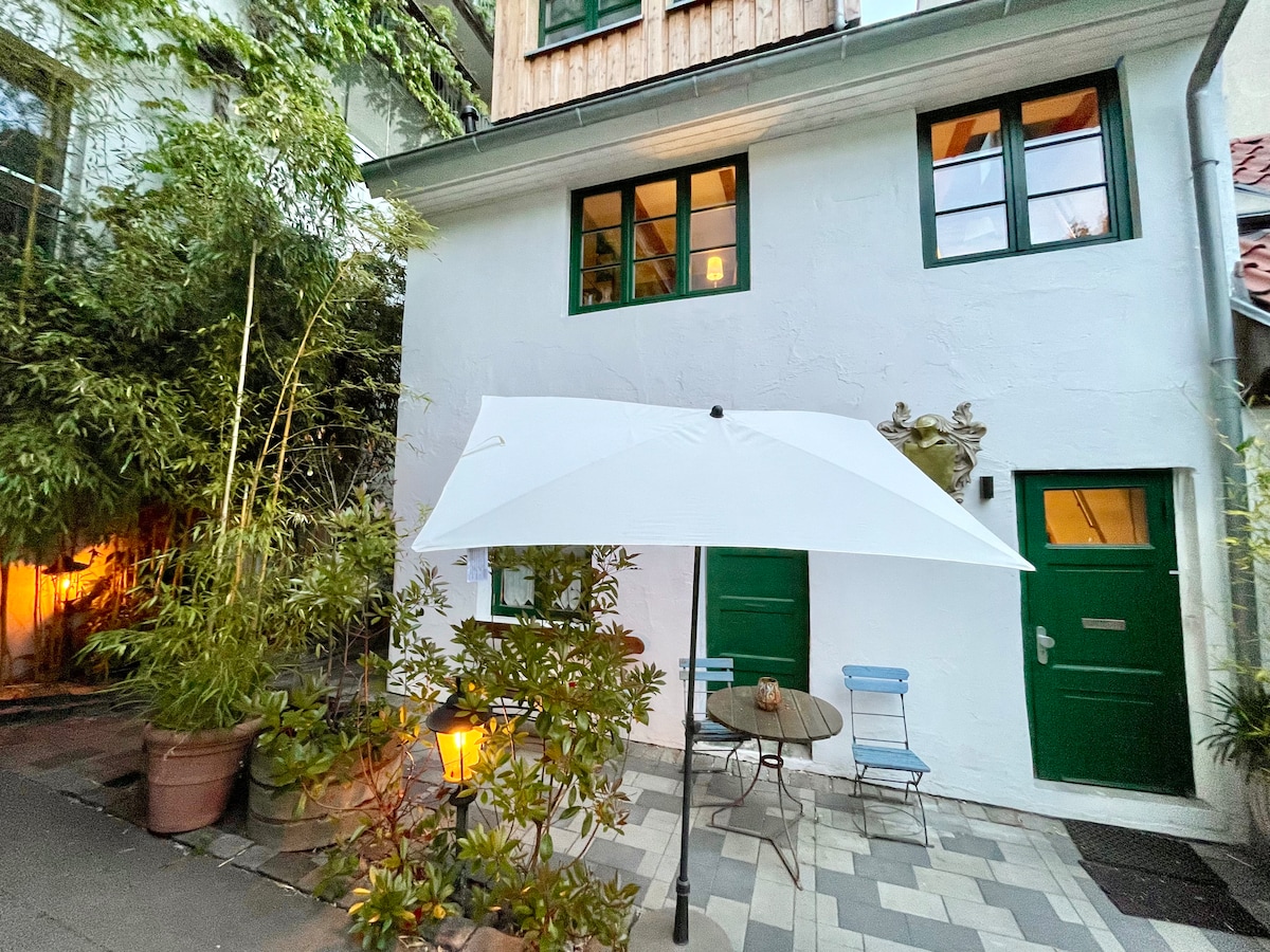 An inviting terrace features a table and two blue chairs under a large white umbrella. Lush greenery surrounds the area, enhancing the serene atmosphere. The charming exterior of the building has green windows and doors, with warm light emanating from plants nearby.