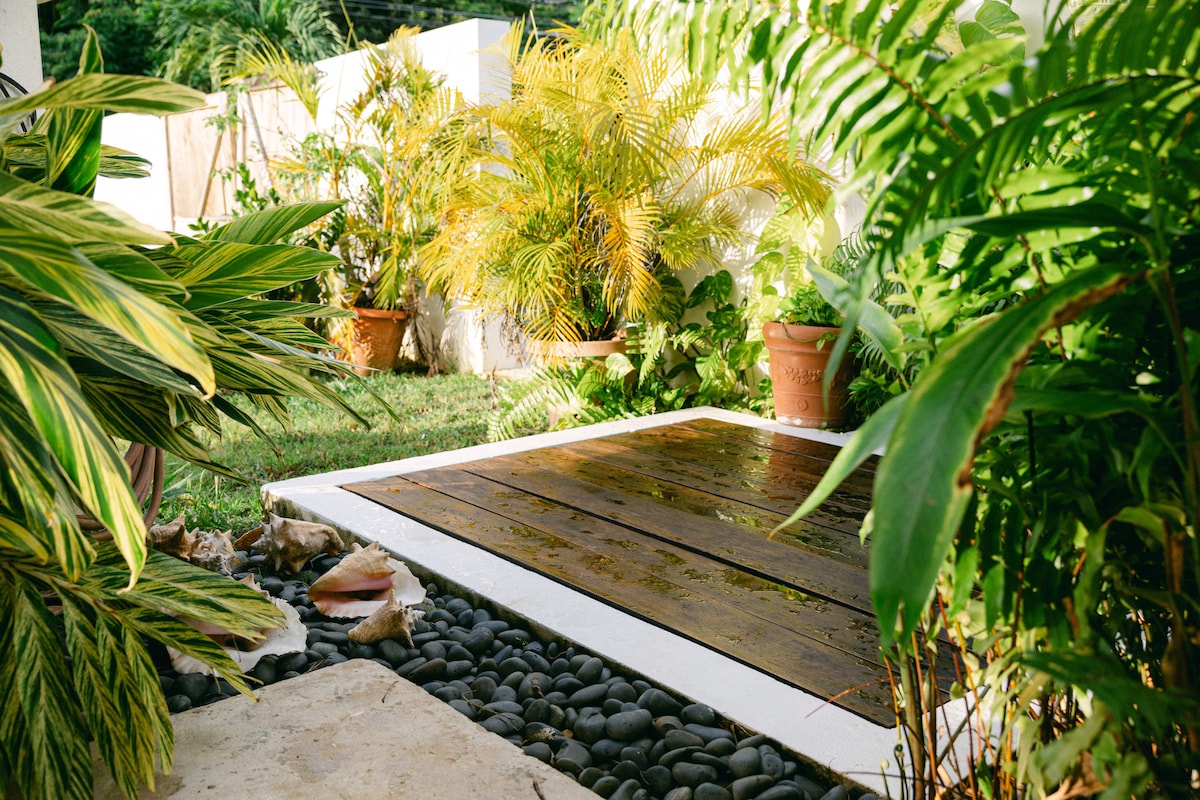 An outdoor space is depicted, featuring a wooden deck surrounded by lush greenery, including potted palms and ornamental plants. Smooth black stones border the deck, enhancing the natural landscaping. The area is clarified by the presence of vibrant foliage, contributing to a serene garden atmosphere.