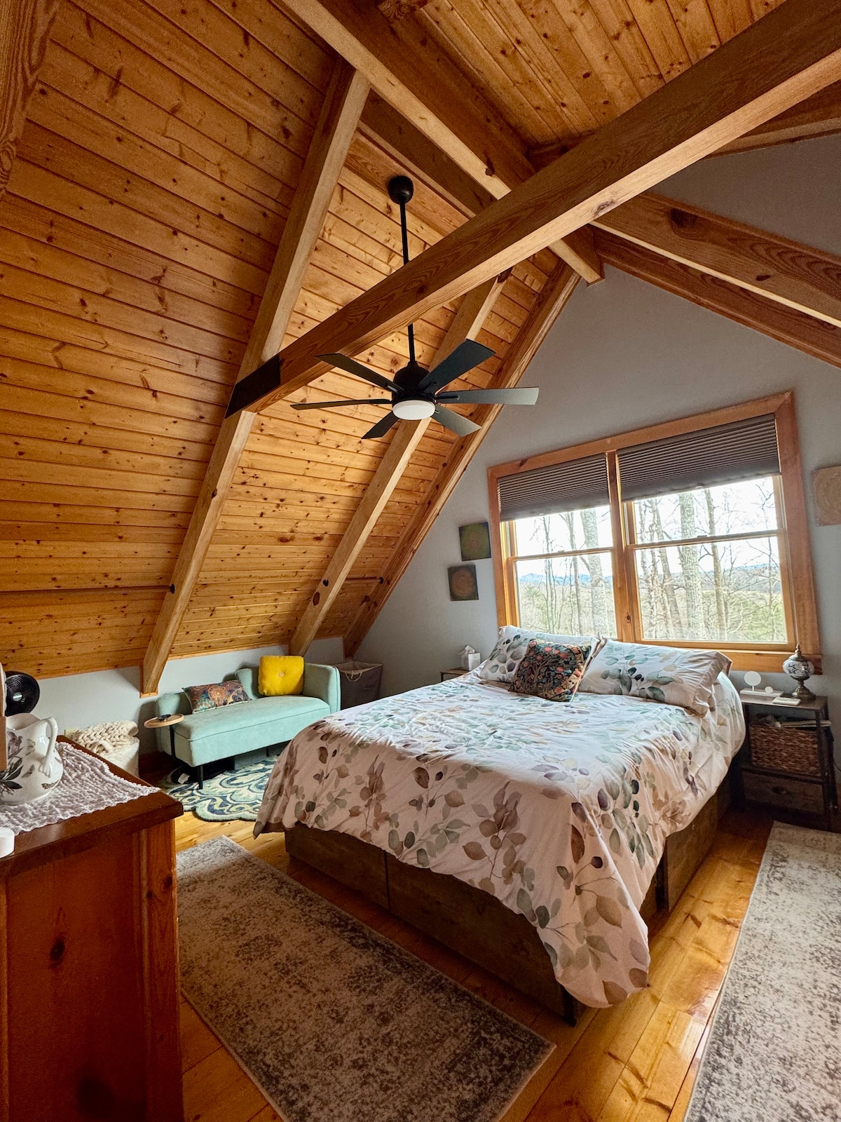 The bedroom features a spacious bed dressed in floral bedding, accompanied by a cozy seating area with natural light streaming through large windows. Exposed wooden beams and a ceiling fan add to the rustic charm, while a soft area rug enhances comfort.