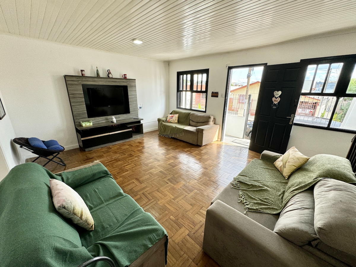 A living room space features two sofas, one draped with a green blanket. A television is mounted on a modern console against one wall. Large windows provide natural light, and a door leads to an exterior balcony area, enhancing the room's openness.